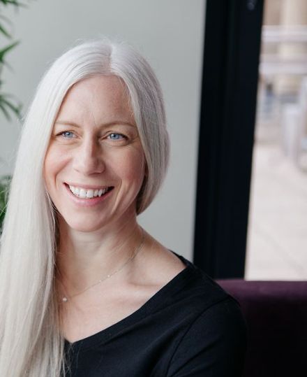 Helen Forsyth smiling, seated by a window, with long silver hair and a black top